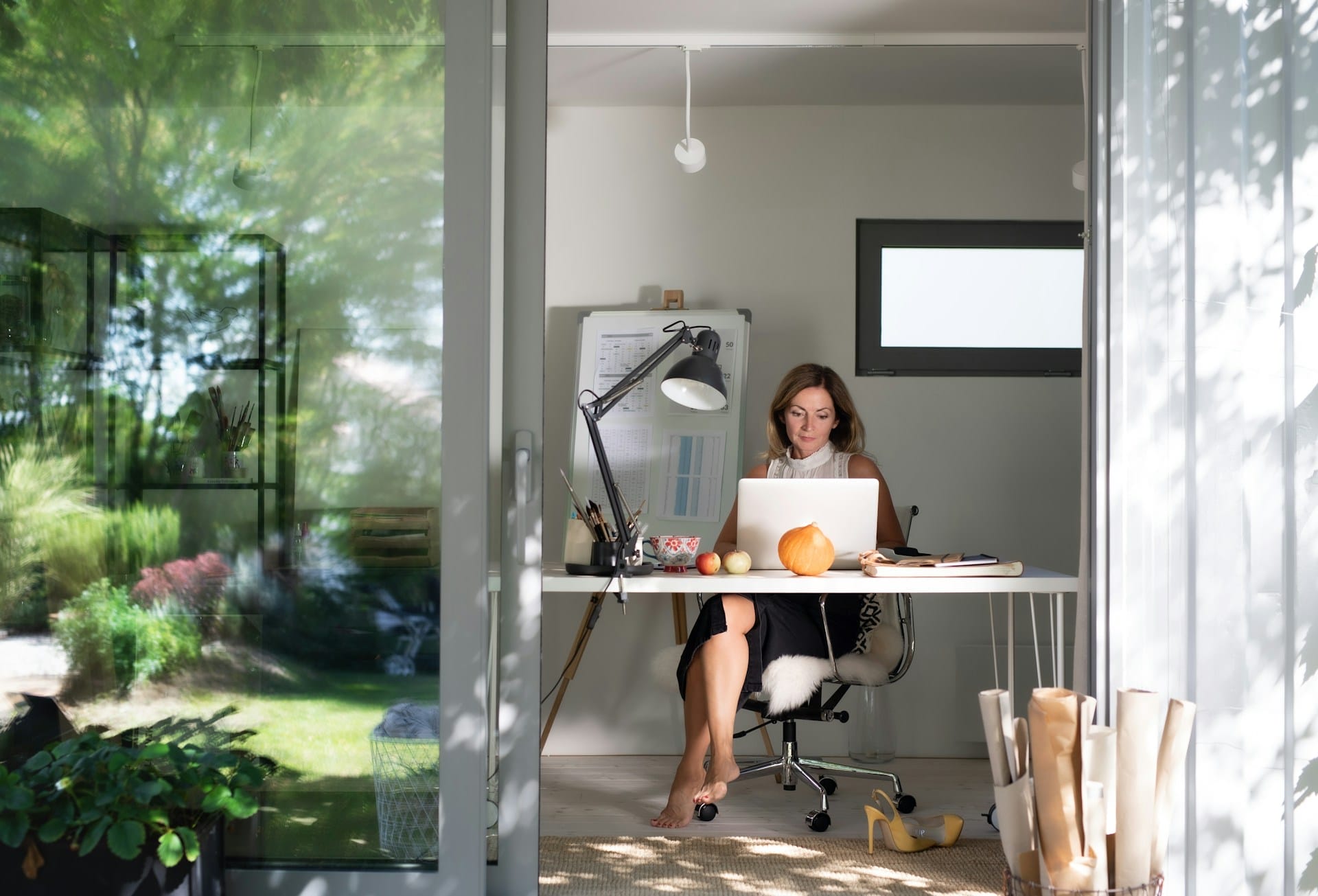 Mature woman working in a bright home office, focused at laptop with natural light and greenery outside.